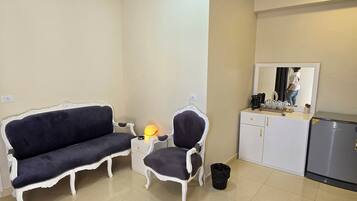 A room with a dark blue sofa, a matching armchair, a small table with a lamp, a white cabinet with a mirror, and a trash bin.