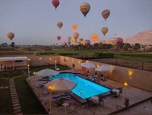 Zona de piscina con sillas de descanso y sombrillas, rodeada de campos y globos aerostáticos al fondo.