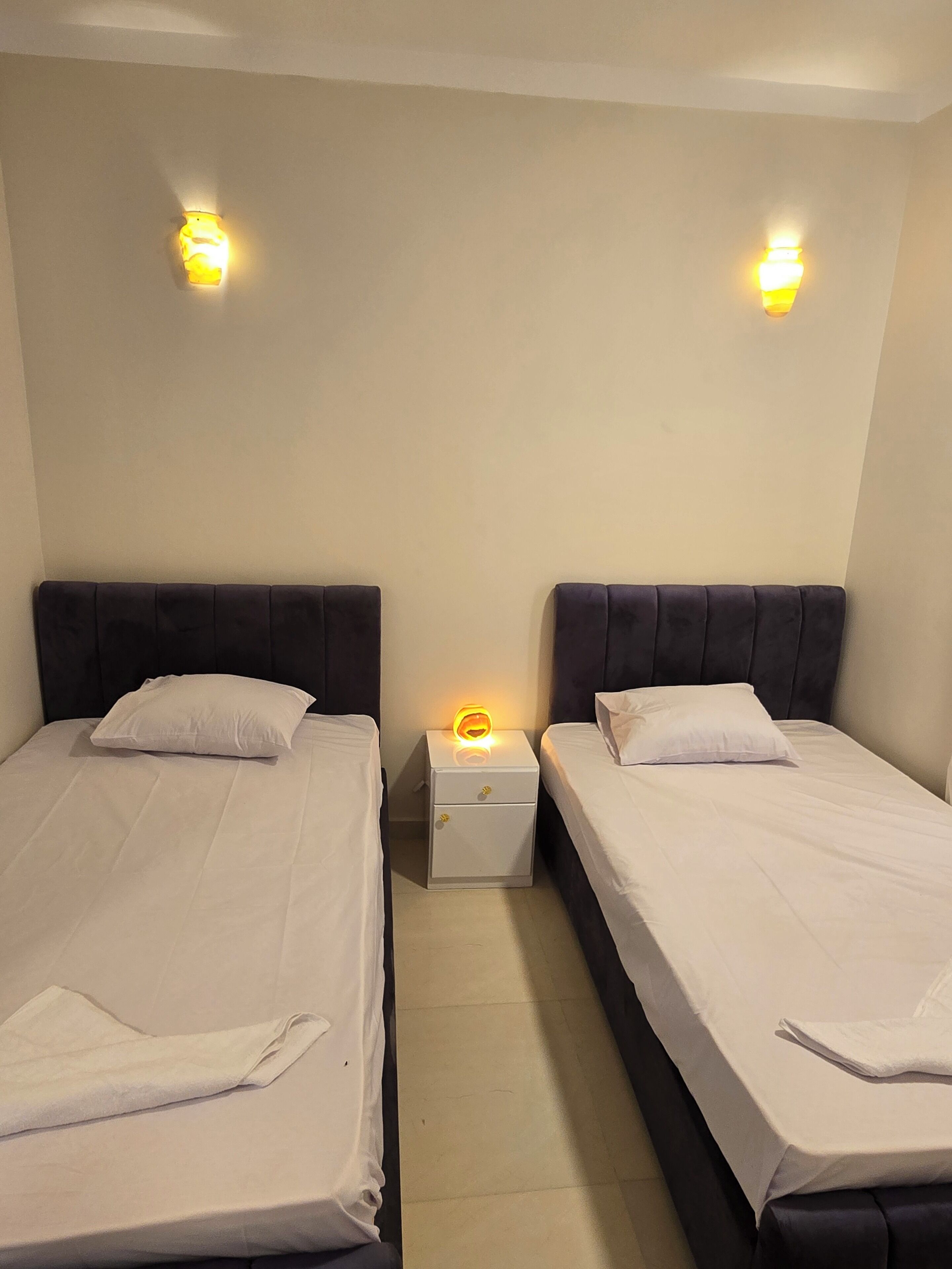 A hotel room with two beds, each with a headboard and a nightstand with a lamp.