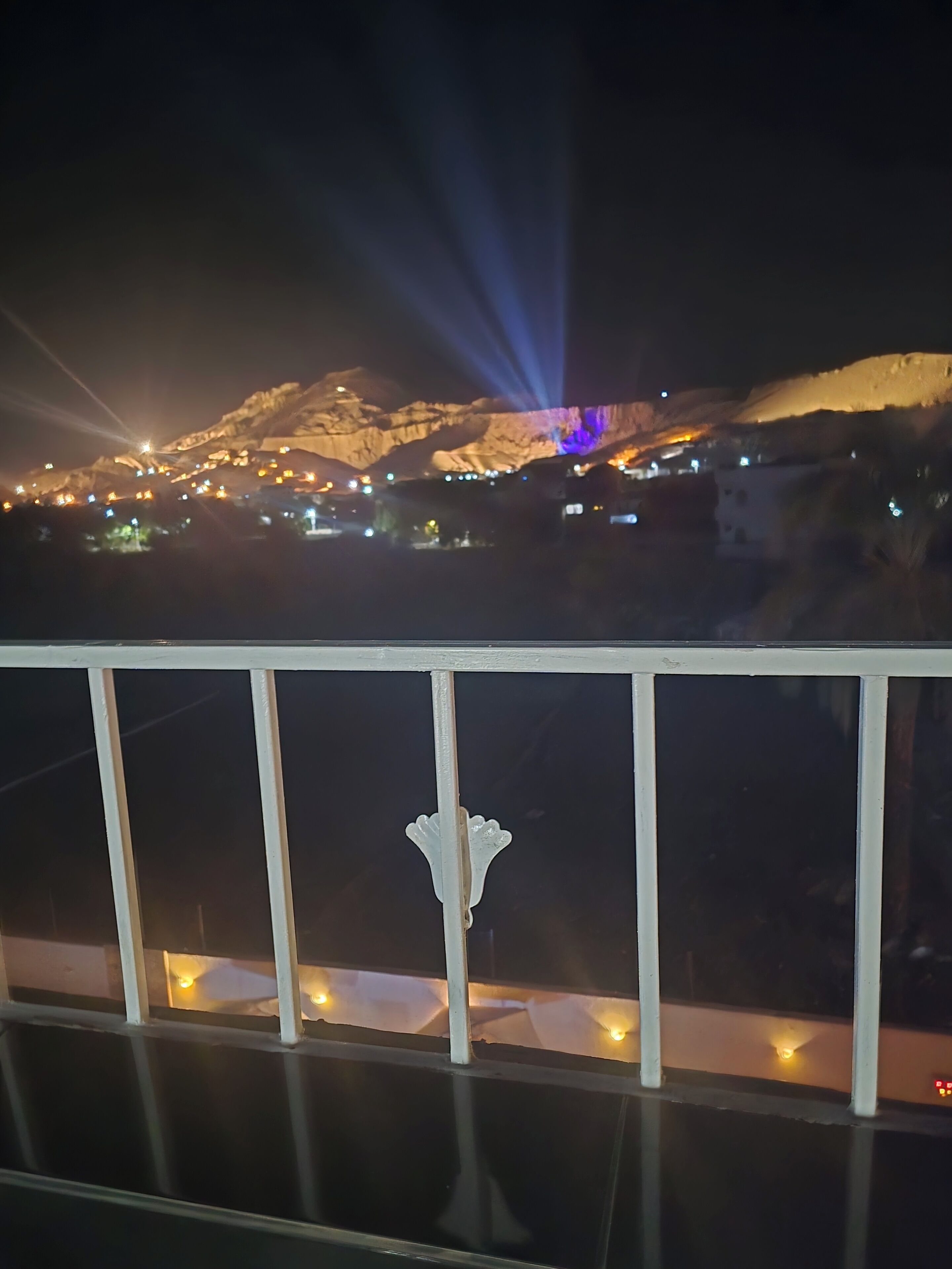 Vista nocturna de una zona montañosa con edificios iluminados y barandilla con luces.