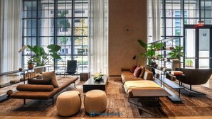 A spacious living room with a sofa set, ottomans, and a coffee table. There are large windows with curtains, a bookshelf, and a plant. - New Luxury Condo in Downtown (Miami)