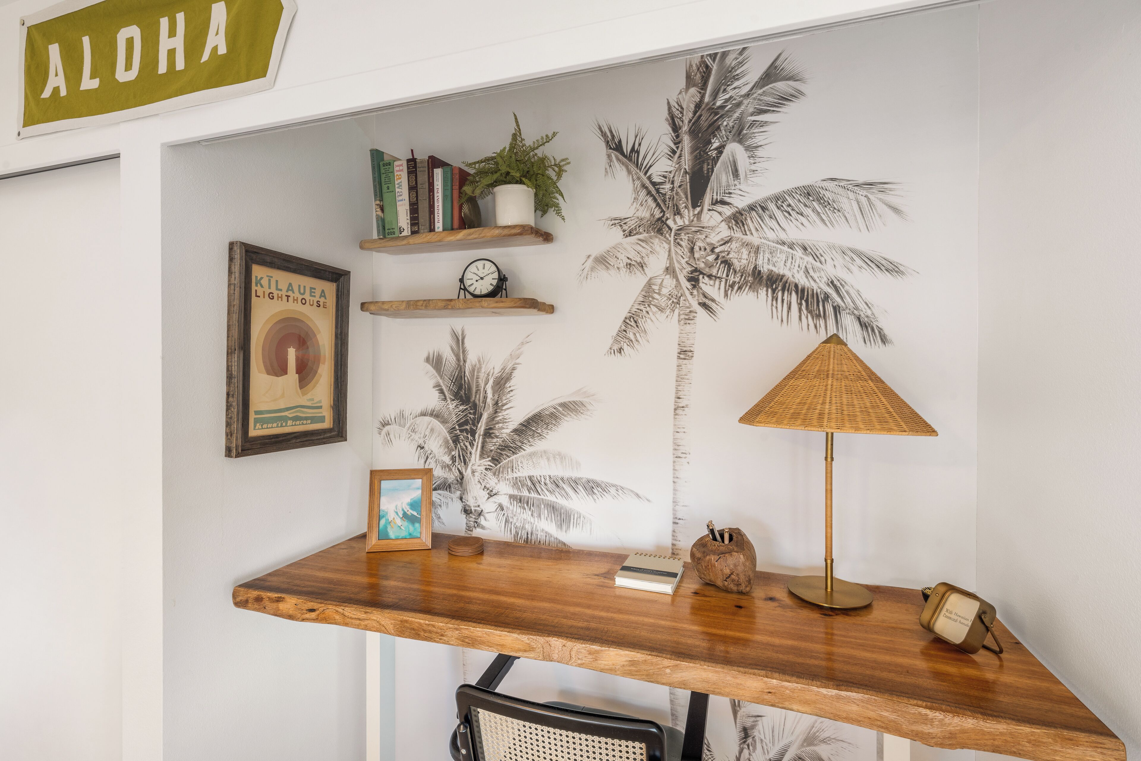 Un espacio de trabajo con un escritorio de madera, un cuadro enmarcado, un reloj, libros y un cuadro mural con motivos de palmera.