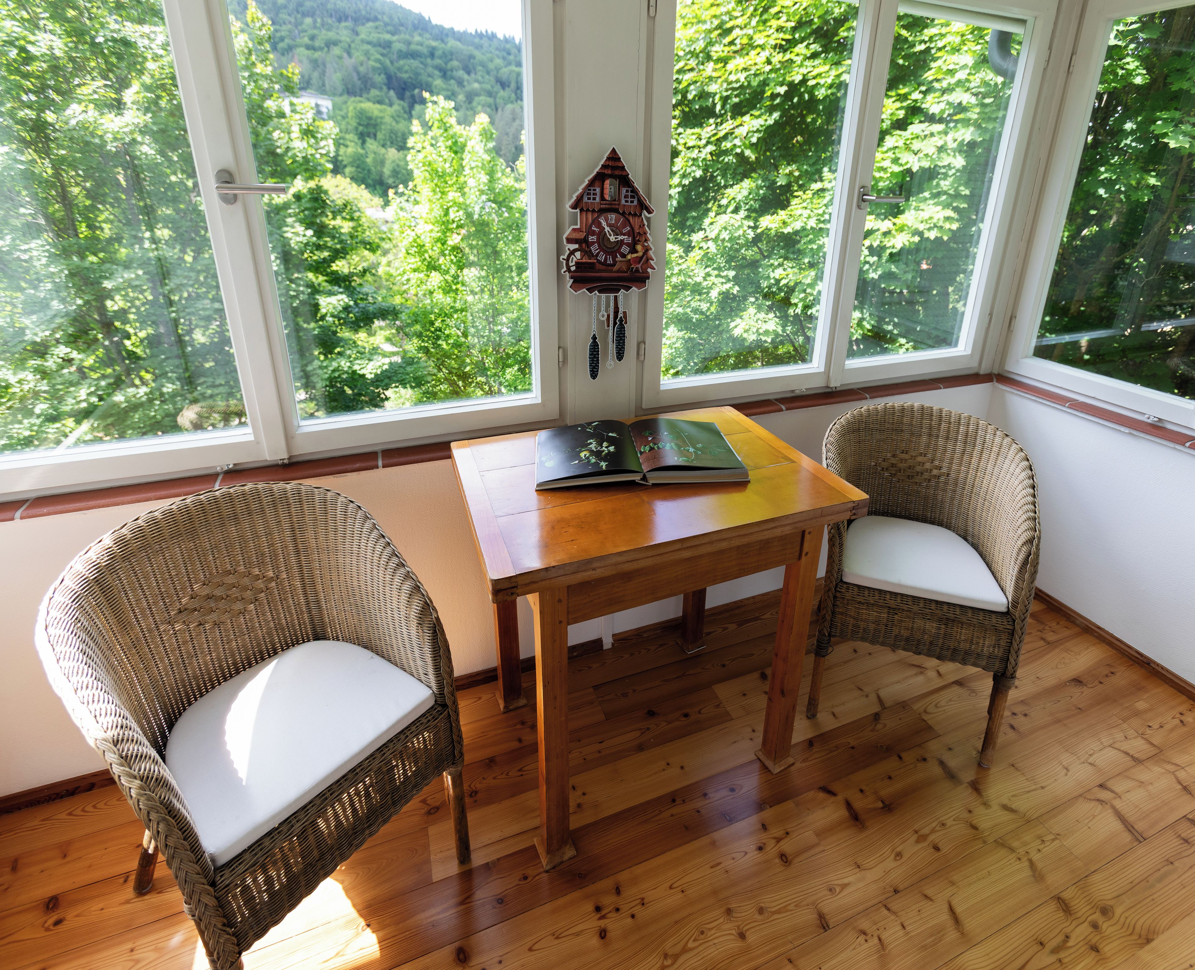 A room with a wooden table, two wicker chairs, a cuckoo clock, and a view of a forest through large windows.