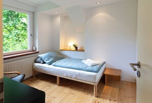 Une chambre à coucher minimaliste, avec un lit simple, une table de chevet, une fenêtre donnant sur un espace verdoyant et un luminaire fixé au mur.
