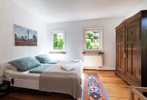 Un dormitorio con una cama, dos ventanas, un radiador, un armario y un cuadro en la pared.