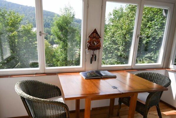 A wooden table with two wicker chairs in a room with large windows overlooking a forested area.