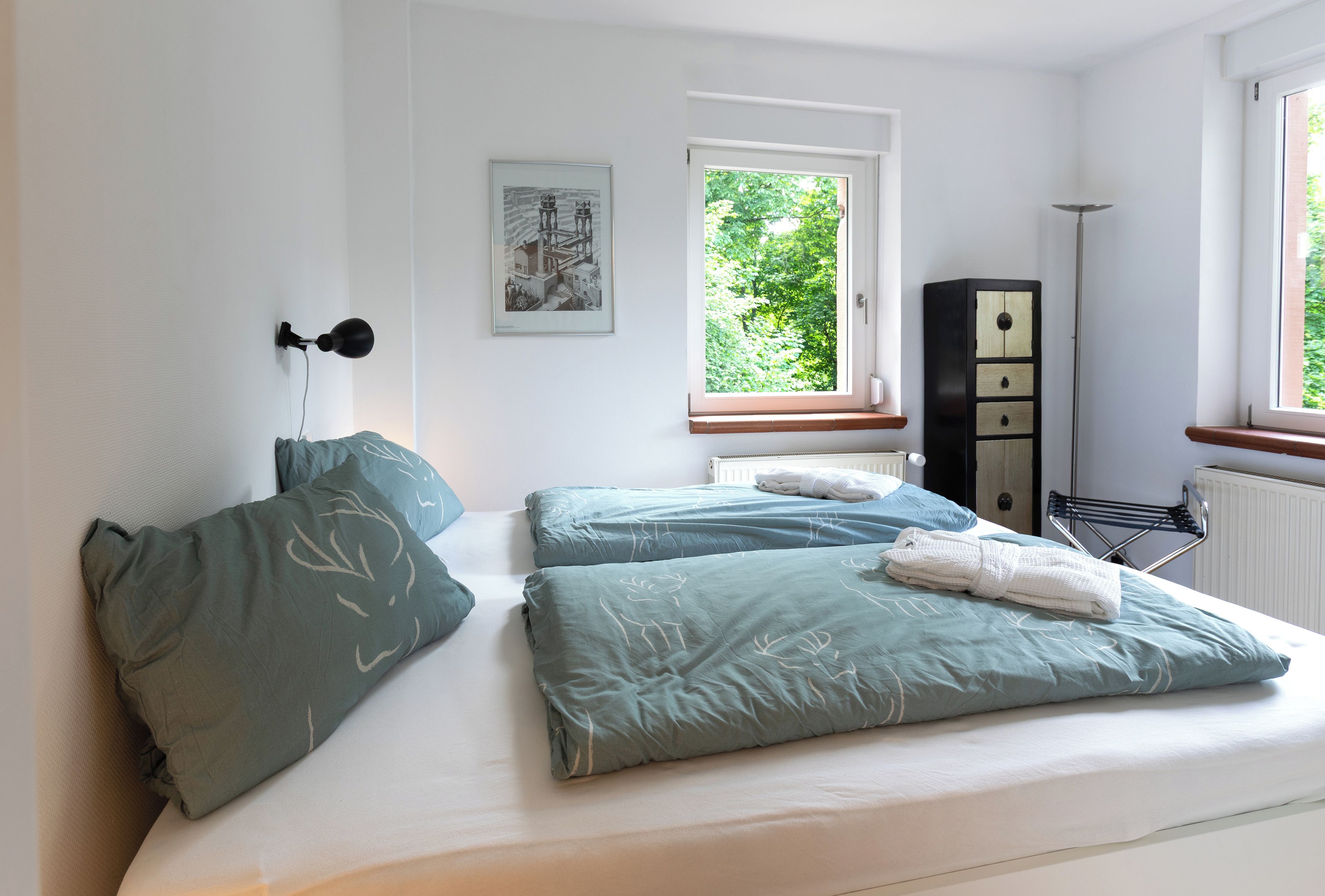 A bedroom with two beds, a window with a view of greenery, a radiator, and a cabinet.