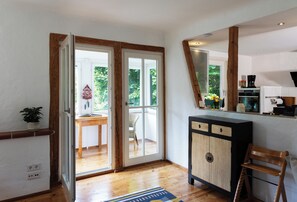 Une pièce avec un plancher en bois, une commode avec des tiroirs, une chaise, une table sur laquelle est posée une plante en pot, et une porte donnant sur l’extérieur.