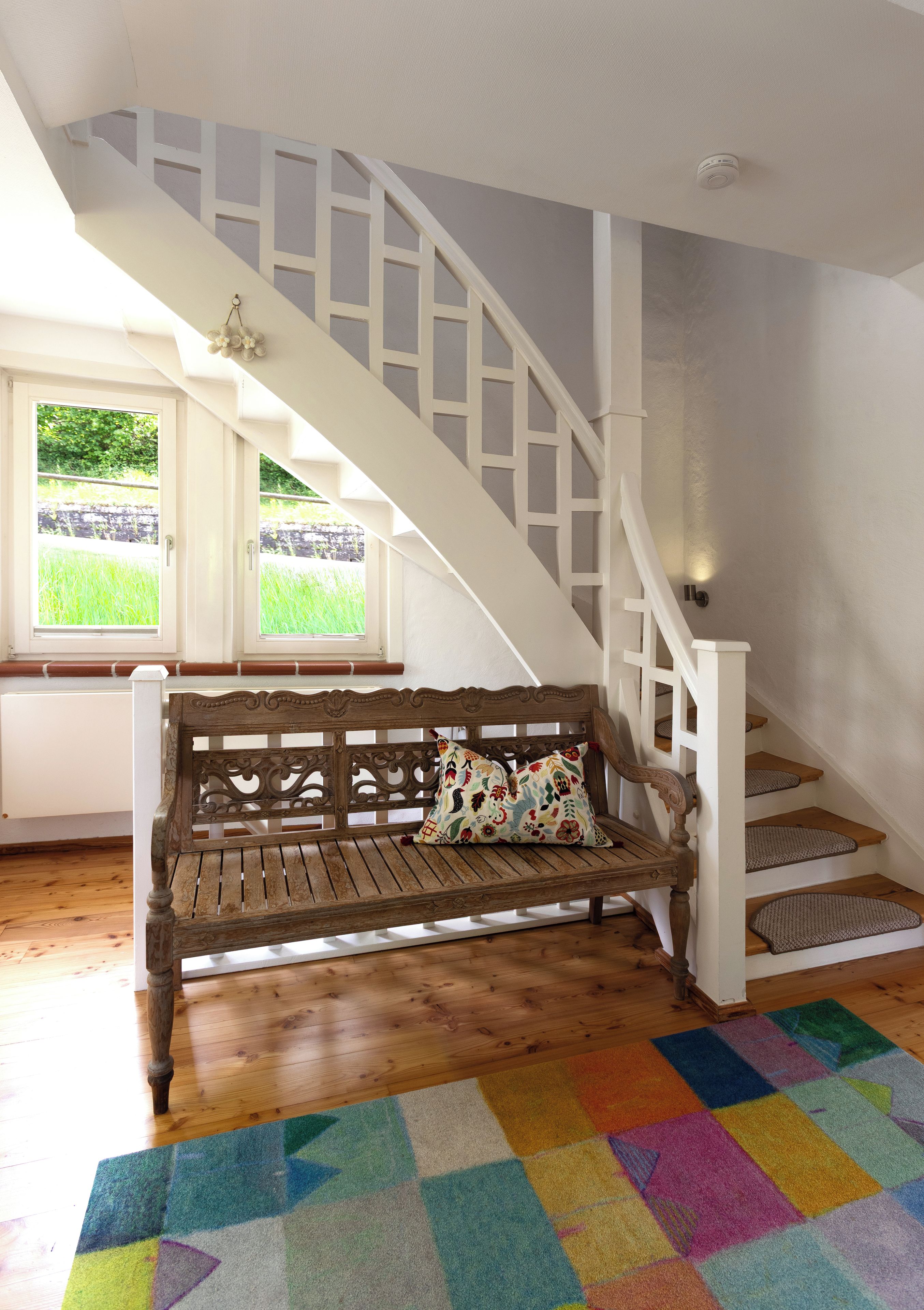 A wooden bench with floral cushions in a room with a white staircase and a colorful rug.