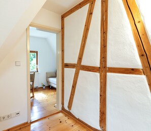 Una habitación con techo inclinado, vigas de madera, pared blanca y suelo de madera.