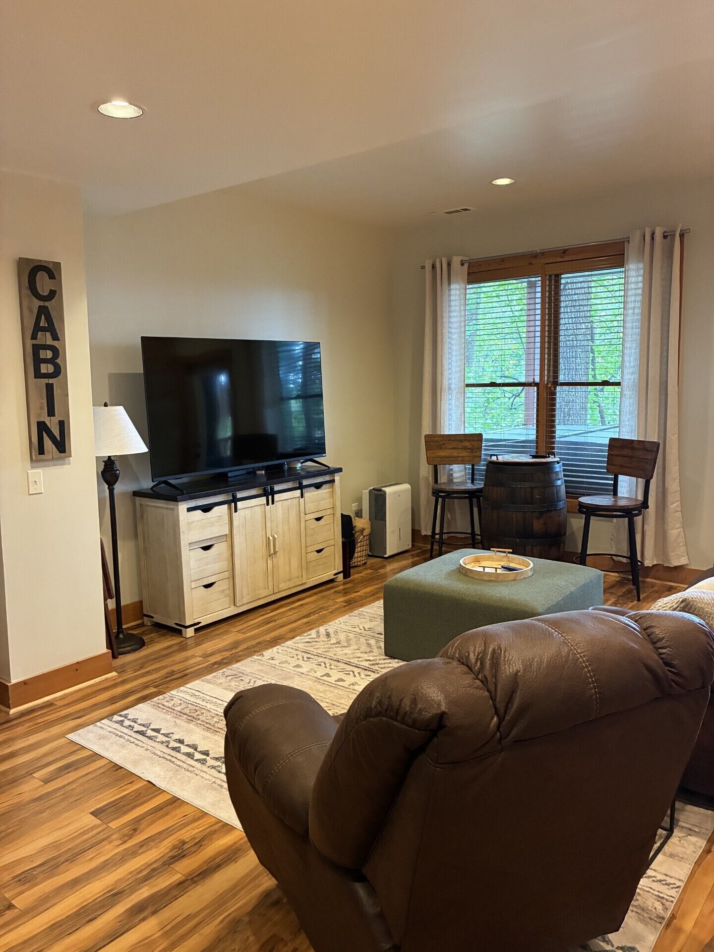 A living room with a flat-screen TV, wooden cabinets, a dining table with chairs, and a large window with curtains.