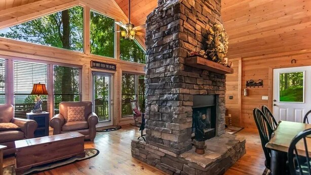 A cozy cabin interior with a stone fireplace, wooden furniture, and large windows offering a view of the outdoors. - Mountain top cabin with big views, a relaxing hot tub and 2 gas fireplaces. (Ellijay)