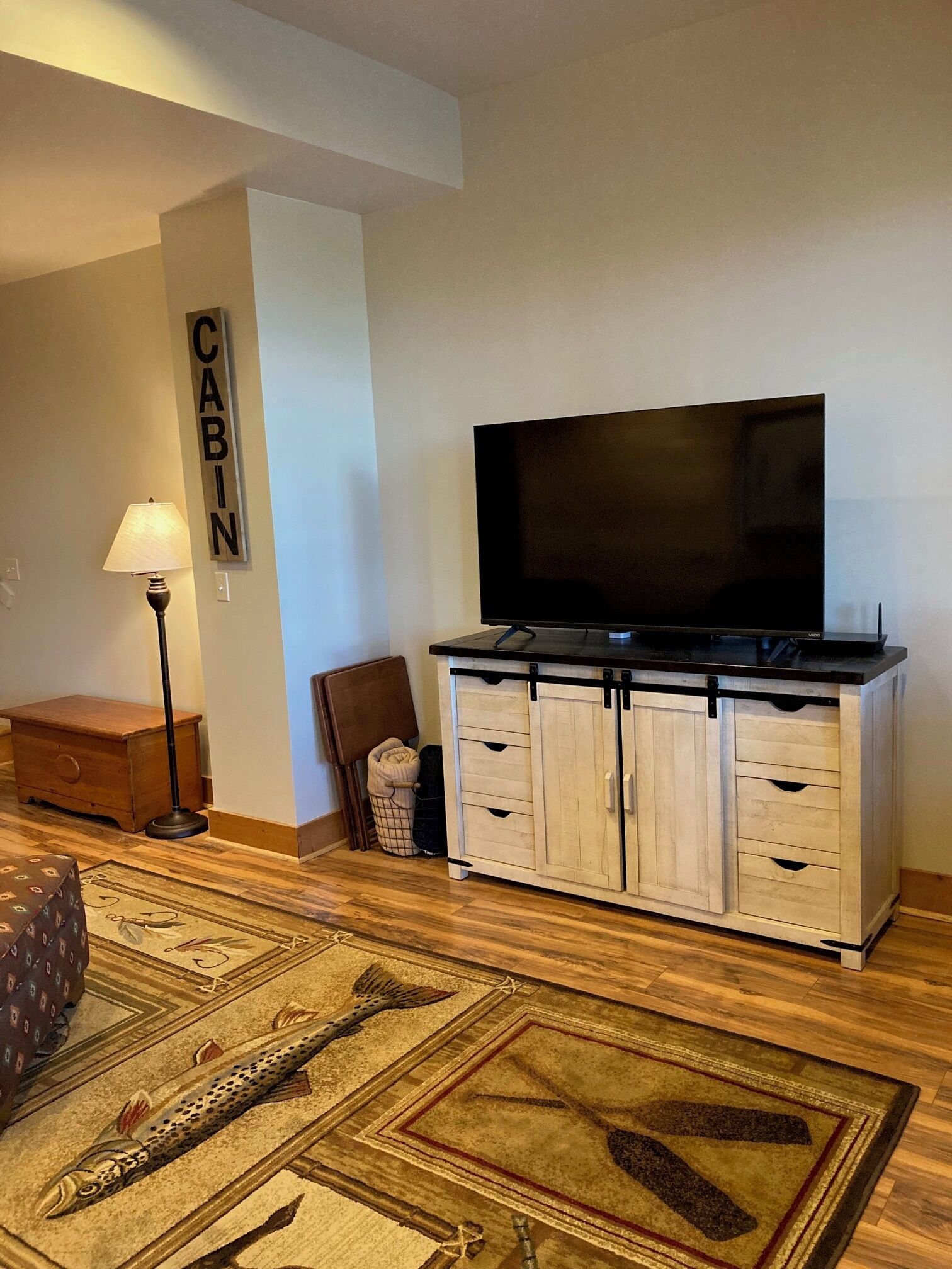 Una sala de estar con un televisor de pantalla plana sobre un mueble de madera, una lámpara, un sillón y un tapete con diseños de peces.