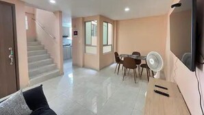A modern living room with a sofa, a dining area with a table and chairs, a staircase, and a flat-screen TV mounted on the wall.