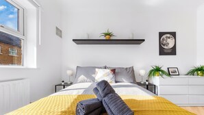 A neatly made bed with a yellow blanket, a grey towel, and a wall-mounted shelf with a plant and a moon poster. - Cozylease 3-Bed Retreat Near O2/LCY with Gym, Workspace, Garden & Free Parking (London)