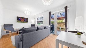 A modern living room with a grey sofa, wooden flooring, a dining table with a plant, and a large window with curtains. - Cozylease 3-Bed Retreat Near O2/LCY with Gym, Workspace, Garden & Free Parking (London)