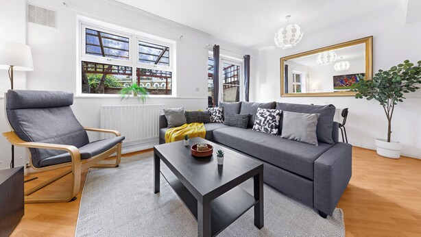 A modern living room with a grey sofa, a wooden chair, a black coffee table, and a large mirror on the wall. - Cozylease 3-Bed Retreat Near O2/LCY with Gym, Workspace, Garden & Free Parking (London)