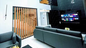 Un hall d’entrée moderne doté d’un grand écran de télévision, de canapés gris, d’une cloison en bois et d’un luminaire au plafond.