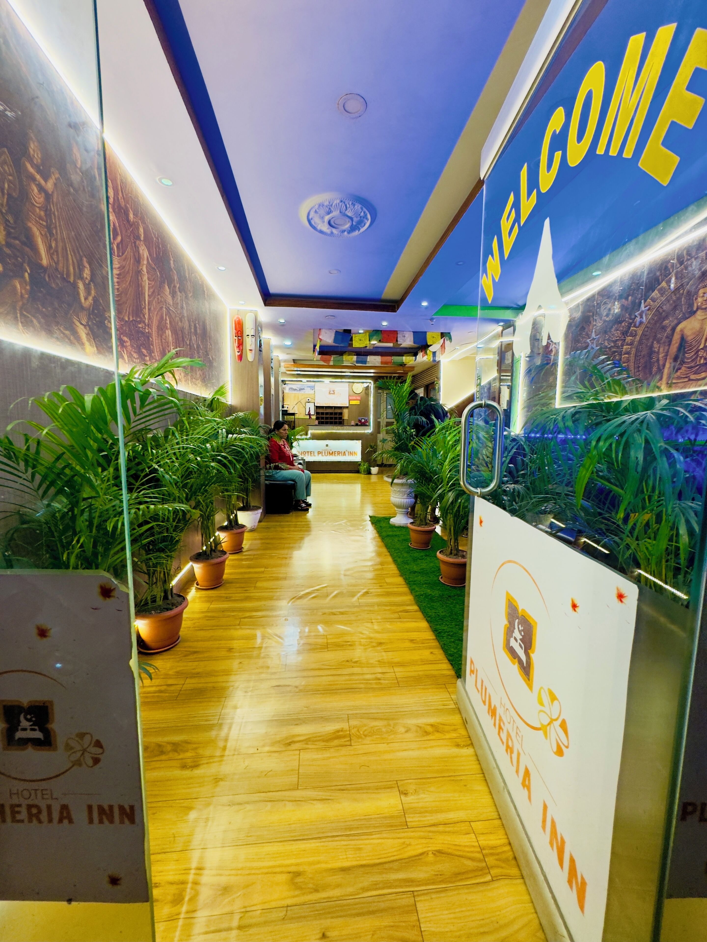A hotel lobby with a wooden floor, potted plants, a reception desk, and a 'Welcome' sign.