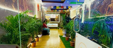 A hotel lobby with a wooden floor, potted plants, a reception desk, and a 'Welcome' sign.