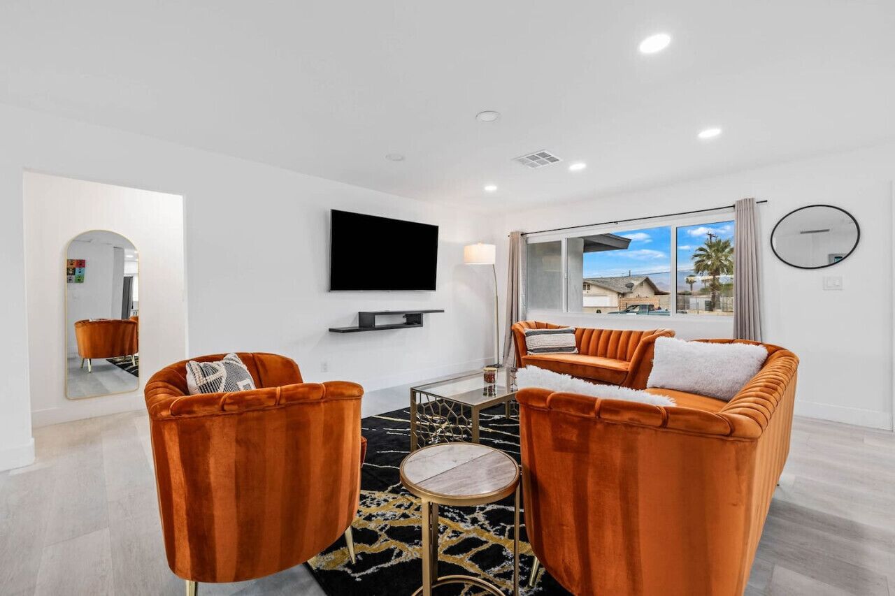 A modern living room with a sofa, two armchairs, a flat-screen TV, and a large window with a view of palm trees.
