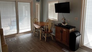 A room with a wooden ceiling, a dining table with chairs, a television mounted on the wall, and a trash can.
