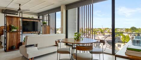 Una sala de estar moderna con un sofá, una mesa de comedor con sillas, un televisor y ventanas amplias con vistas a la ciudad.