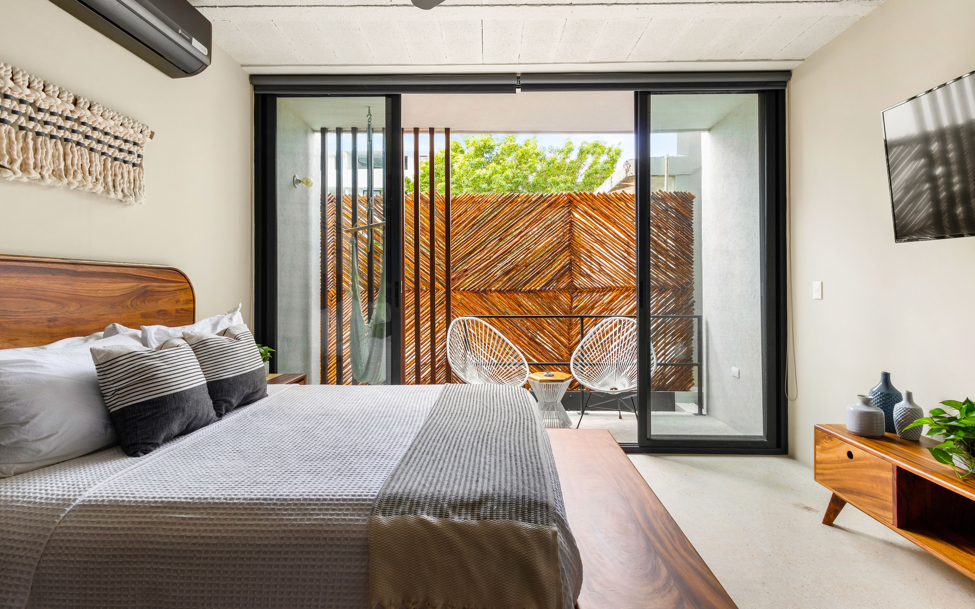 A modern bedroom with a large bed, a wooden headboard, a sliding glass door leading to an outdoor area with a woven fence, and a wooden side table with a vase.