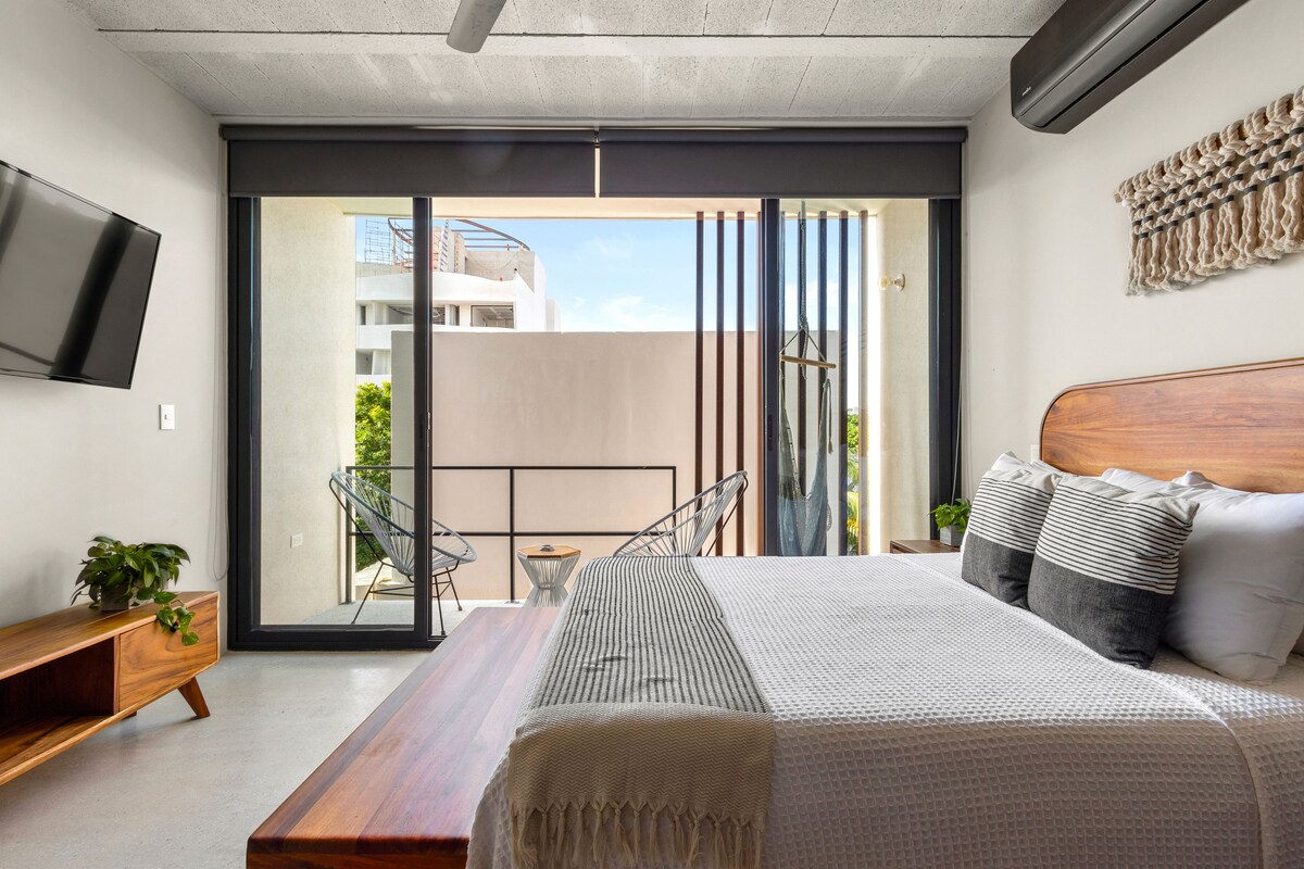 A modern bedroom with a large bed, a TV, and a balcony with a view of buildings.