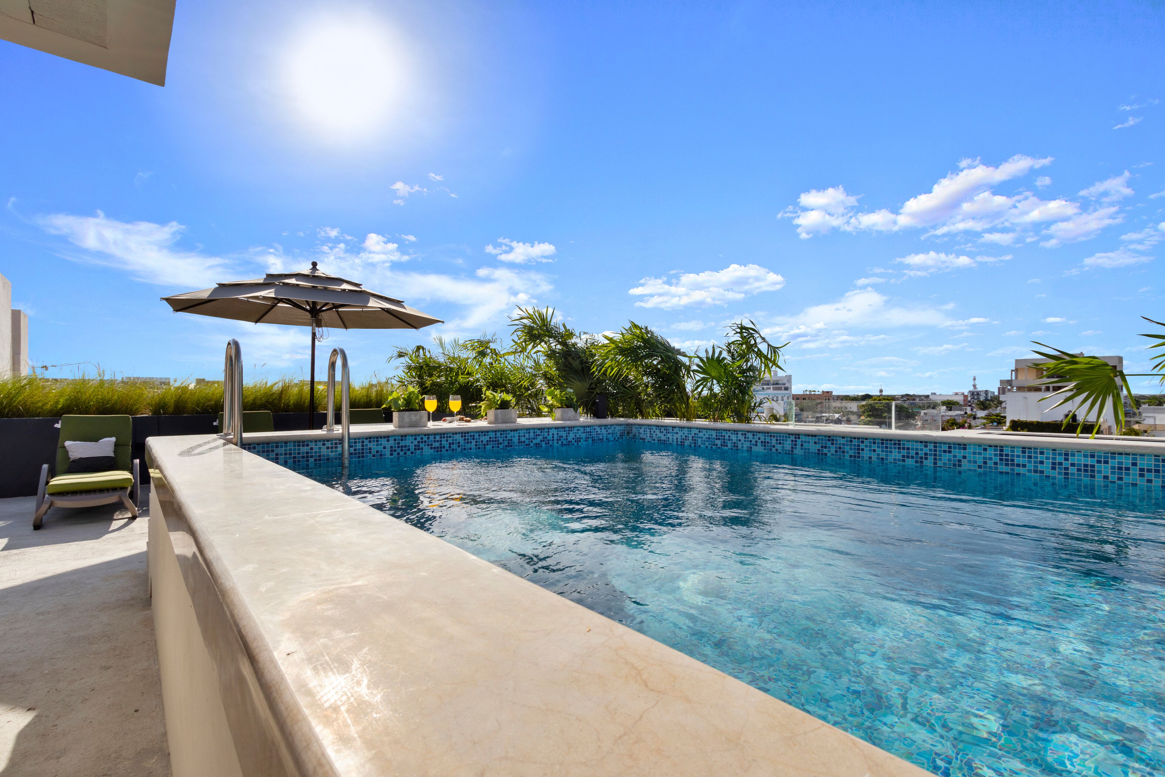 Une piscine, un ciel bleu azur et quelques nuages. On aperçoit une chaise longue et un parasol près de la piscine.