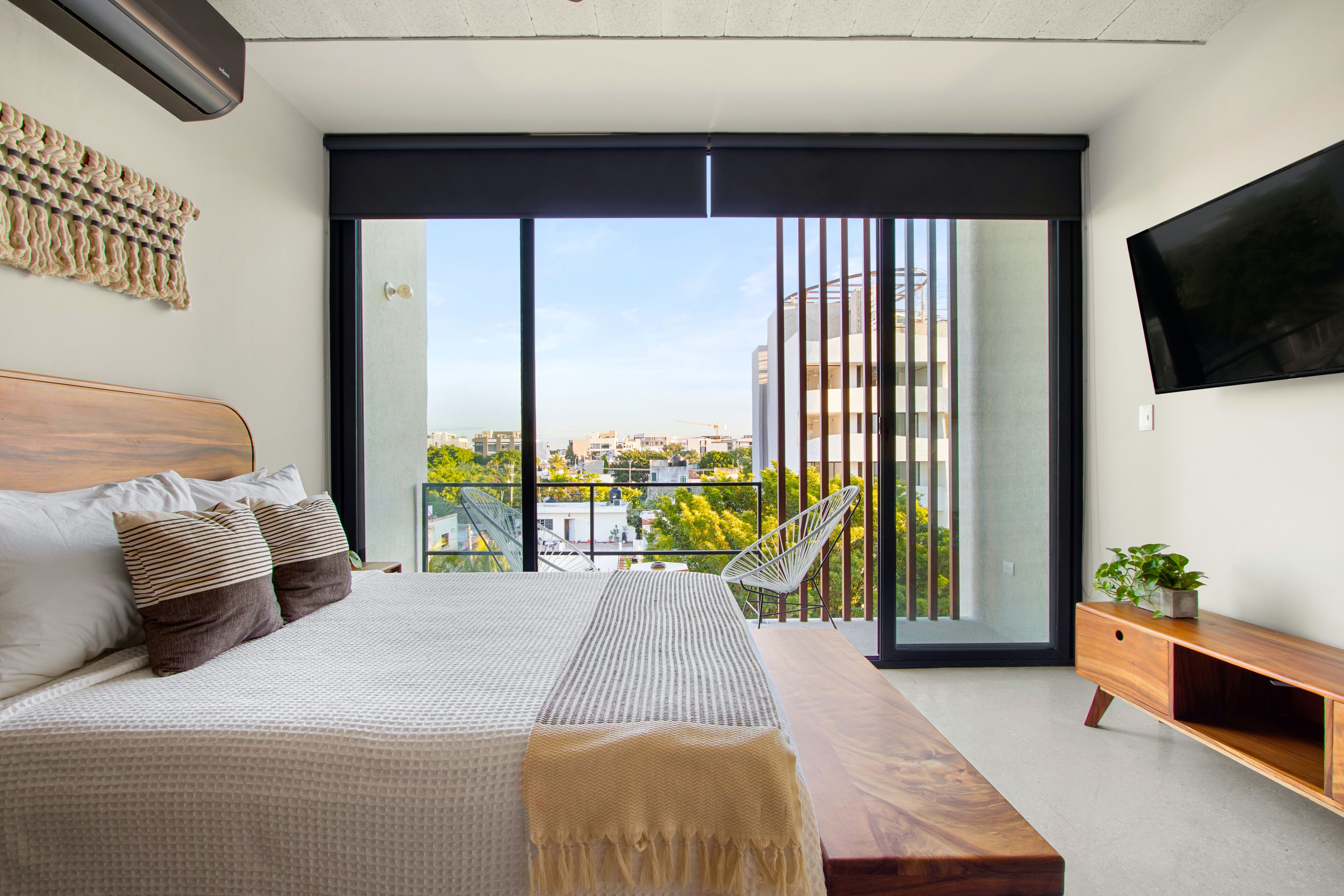 Un dormitorio moderno con una cama grande, un banco de madera, un televisor y un balcón con vistas a edificios y árboles.