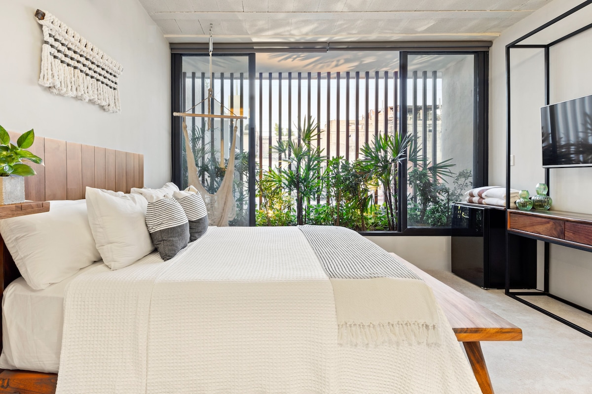 A modern bedroom with a large bed, a wooden headboard, a macrame wall hanging, and a view of greenery through a window with vertical blinds.