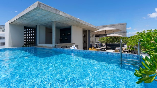 Piscina con sillas de descanso y sombrillas, un área de estar cubierta y un edificio moderno con techo de hormigón.
