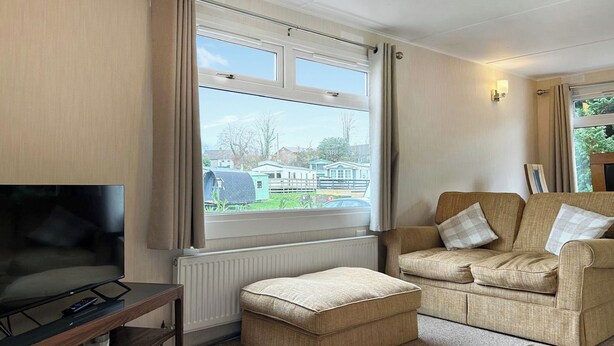 A living room with a TV on a wooden stand, a beige sofa with cushions, a small ottoman, and a window with curtains. - 3 bedroom accommodation in Newton Stewart (Newton Stewart)