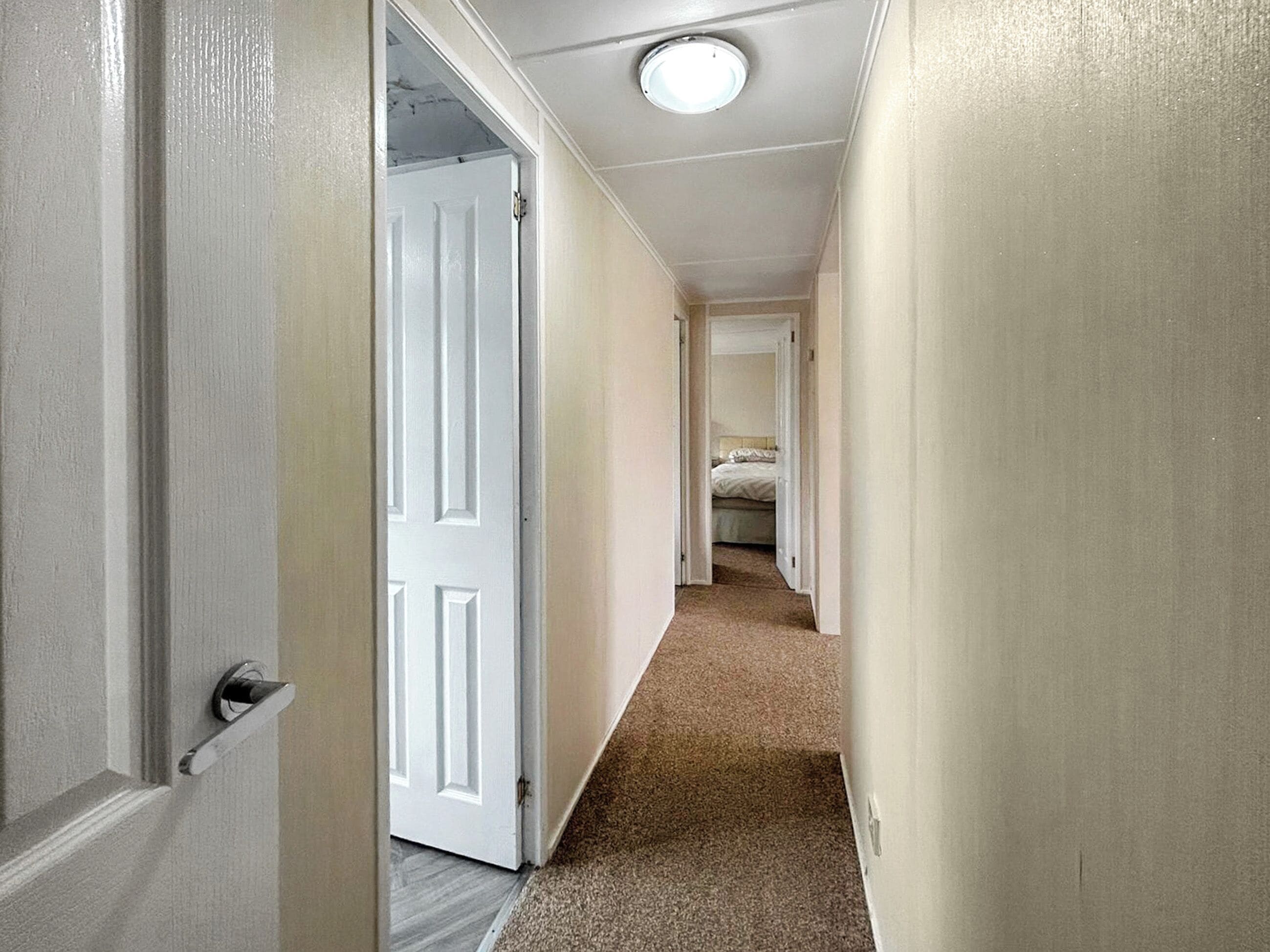 Un couloir étroit aux murs blancs et au sol recouvert d’une moquette, qui mène à une chambre à coucher avec un lit et une table de chevet.