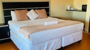Un lit bien fait, avec une couette blanche et un plaid beige, une tête de lit noire et une table de chevet sur laquelle se trouvent une bouilloire et une tasse.