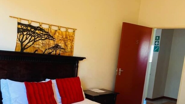 Un lit bien fait, avec du linge de lit blanc et rouge, une tête de lit en bois, une porte rouge et un mur orné d’une tapisserie représentant un arbre.