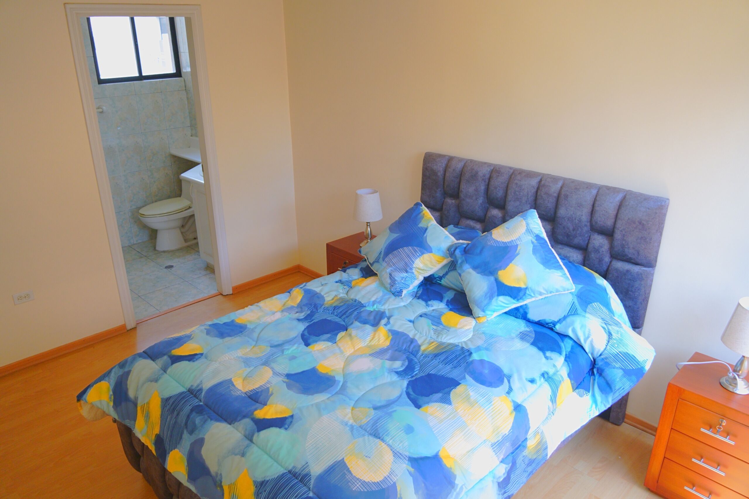 Un dormitorio con cama, mesita de noche, lámpara y vista al baño.