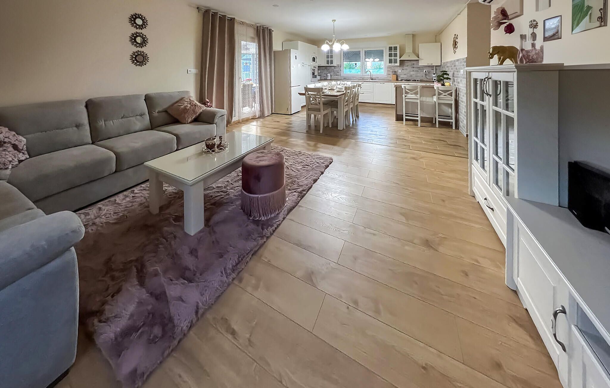 A modern living room with a grey sofa, a white coffee table, and a purple ottoman. A kitchen with white cabinets and a dining area with a table and chairs is visible in the background.