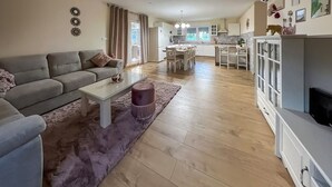A modern living room with a grey sofa, a white coffee table, and a purple ottoman. A kitchen with white cabinets and a dining area with a table and chairs is visible in the background. - Awesome home in Krapinske Toplice (Krapinske Toplice)