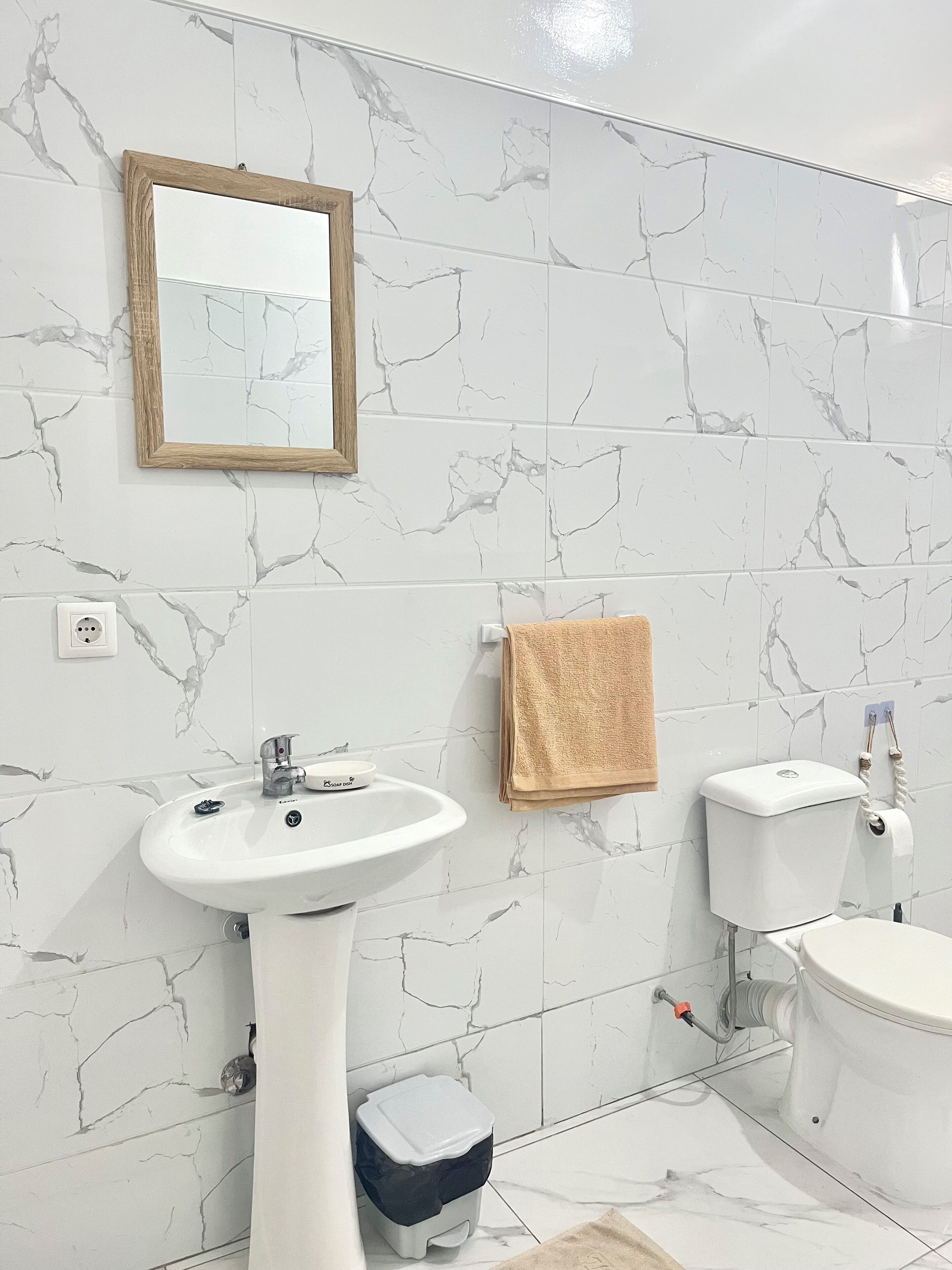Et badeværelse med hvide marmortile, en hvid vask på fod, toilet, en lille skraldespand, et håndklæde hængende på væggen og et indrammet spejl.
