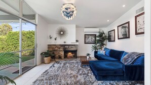 A modern living room with a blue sofa, a fireplace, and a large window overlooking greenery. - Mid Century Modern home -newly renovated with swim jet pool, spa on golf course! (Palm Desert)