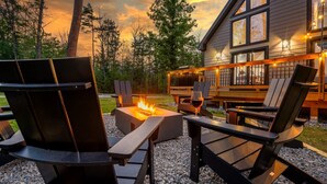 A cozy outdoor seating area with Adirondack chairs and a fire pit, surrounded by a wooden deck and a house with large windows. - Large Trendy Chalet, HotTub, Sleeps 14, Arcade! (Bethel)