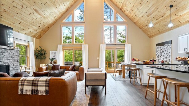 A spacious loft-style living area with a stone fireplace, a large sofa, and a kitchen with bar stools. - Large Trendy Chalet, HotTub, Sleeps 14, Arcade! (Bethel)