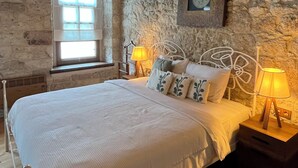 Une chambre avec un plafond en bois, des murs en pierre, un lit avec une literie blanche, deux lampes de chevet, une fenêtre et une horloge au mur.