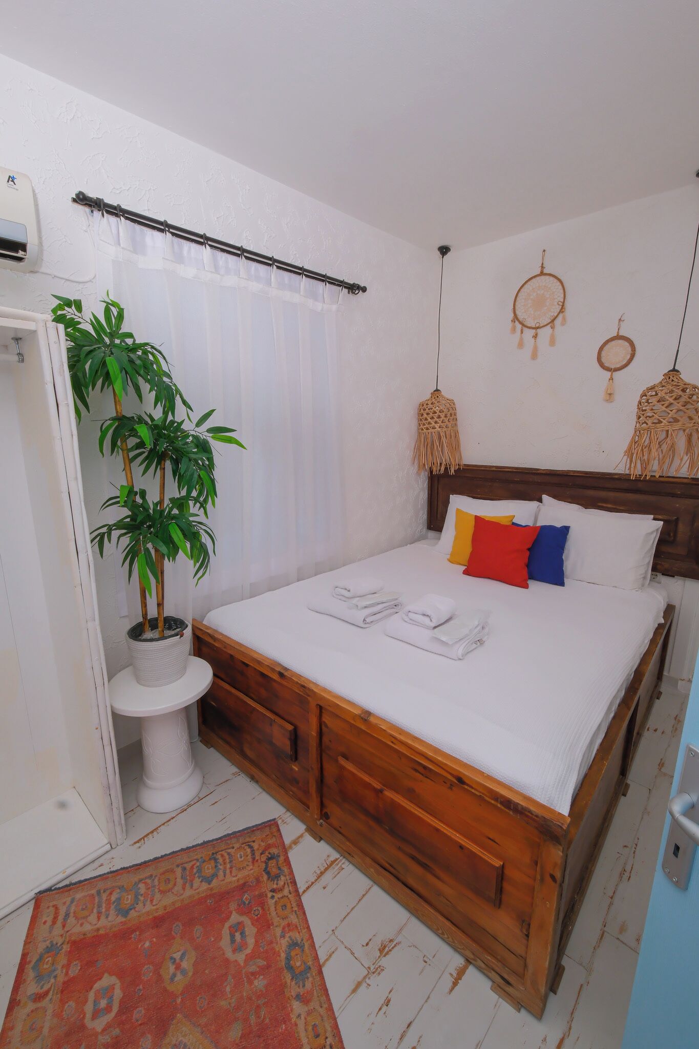 Un dormitorio con una cama de madera, ropa de cama blanca, una planta en maceta, un inodoro blanco y una alfombra estampada.