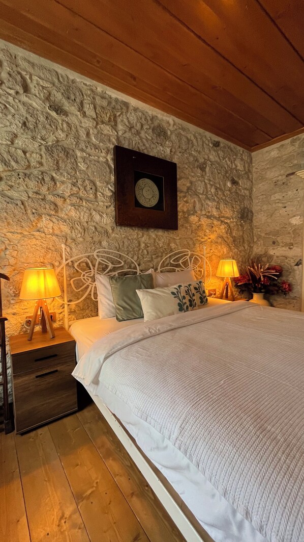 A bedroom with a stone wall, a bed with white bedding, two bedside tables with lamps, and a wooden floor. - Sisu Alacati (Cesme)