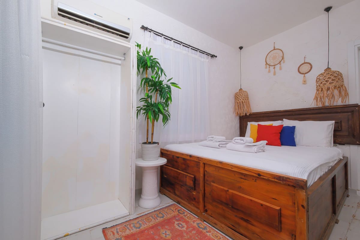 A bedroom with a wooden bed, white bedding, a potted plant, and a small white pedestal table.