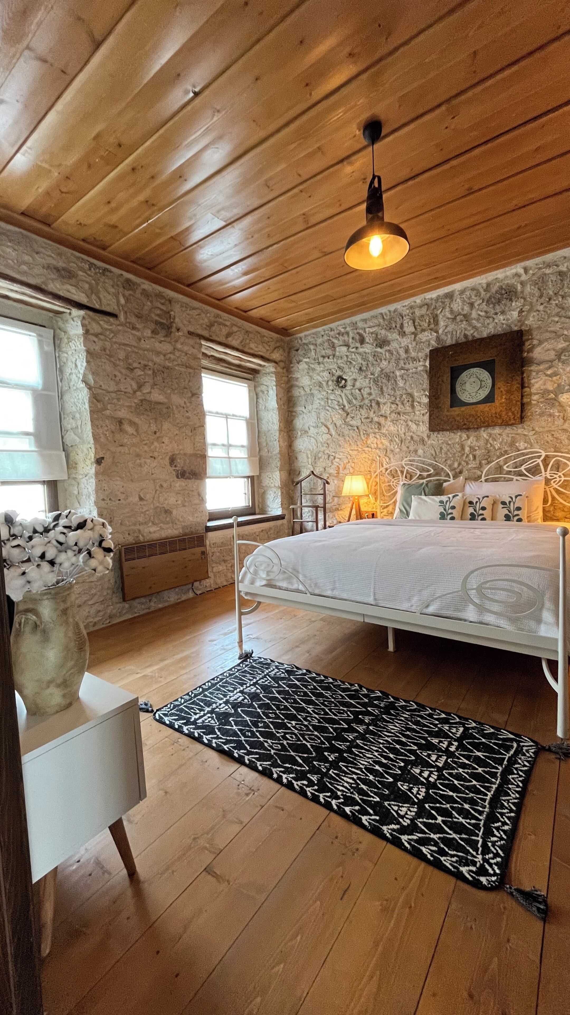 A bedroom with a stone wall, wooden ceiling, a bed with a white headboard, a black and white rug, and a window with curtains.