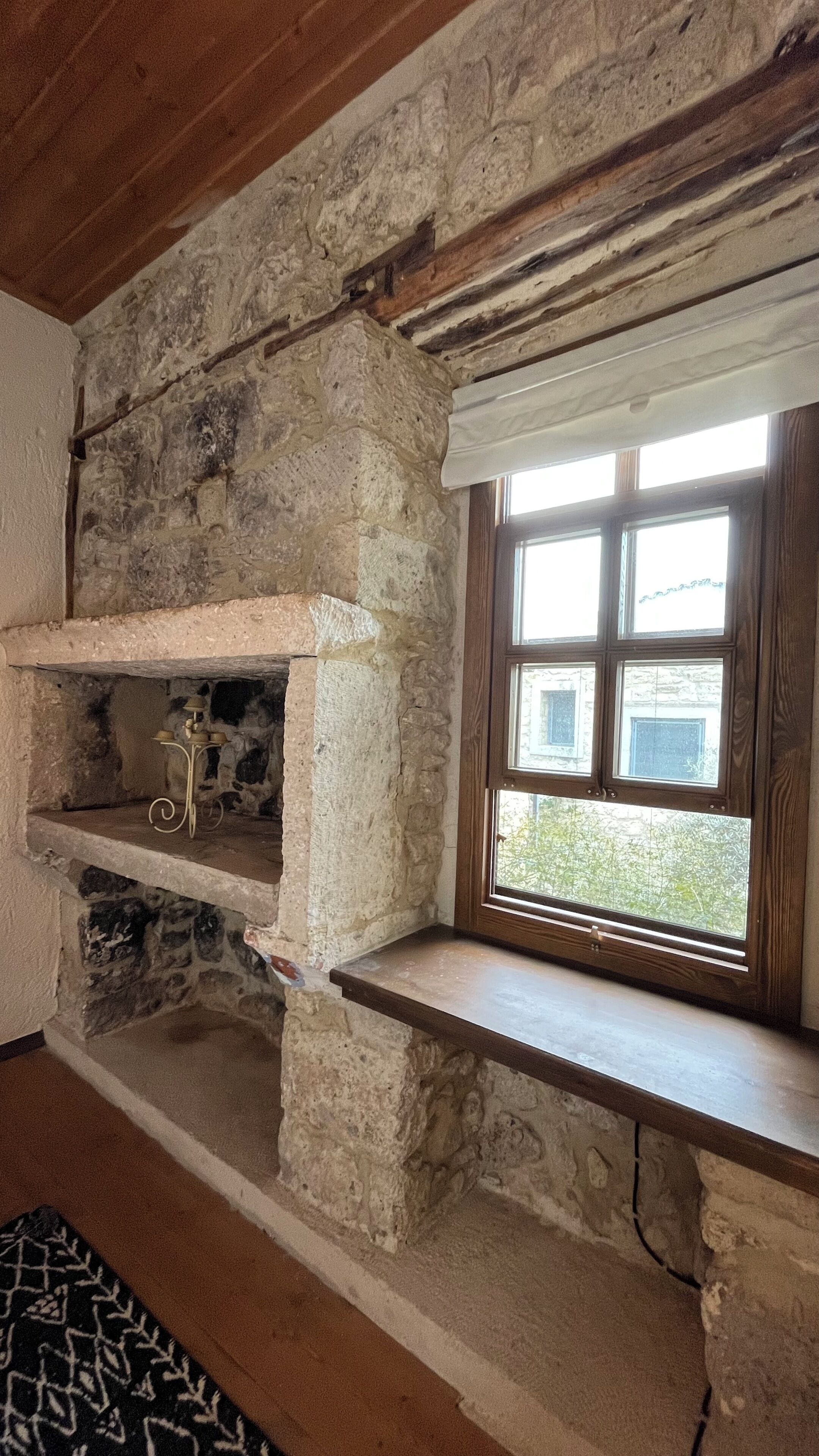 A stone fireplace with a wooden shelf, a window with a view of greenery, and a patterned rug on the floor.